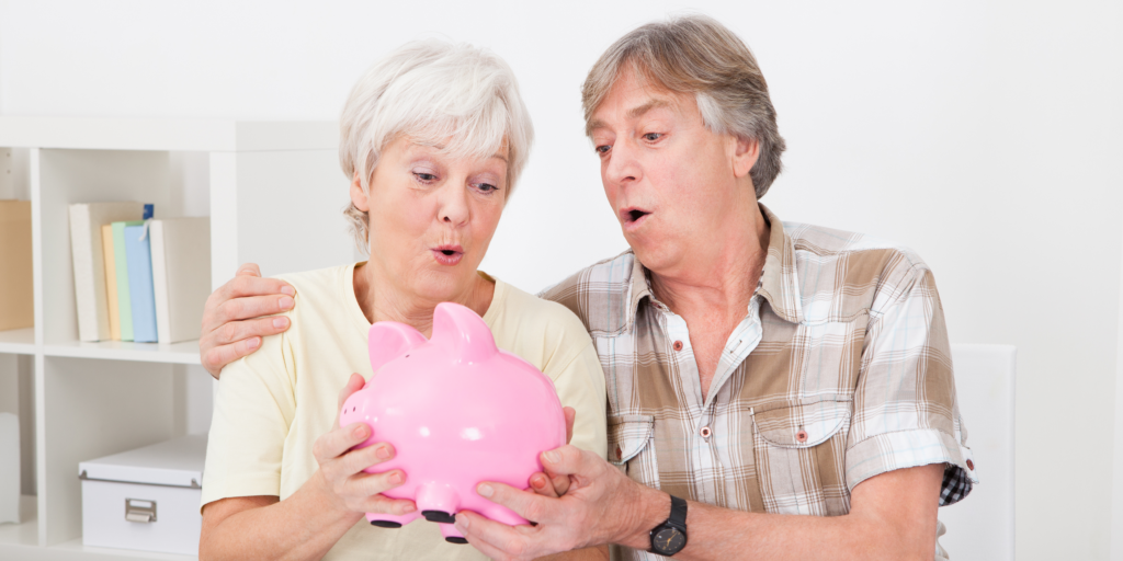 An elderly couple with light skin, sitting together in a bright living room, holding a pink piggy bank. They both have excited and surprised expressions as they look at the piggy bank, symbolizing savings or financial planning. The woman has short white hair and is wearing a light yellow shirt, while the man has gray hair and is wearing a plaid button-up shirt.