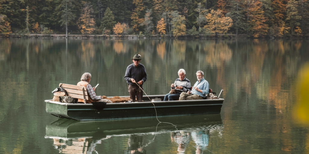 Four elderly men fishing from a small boat on a calm lake surrounded by autumn trees, with colorful foliage reflected in the water.